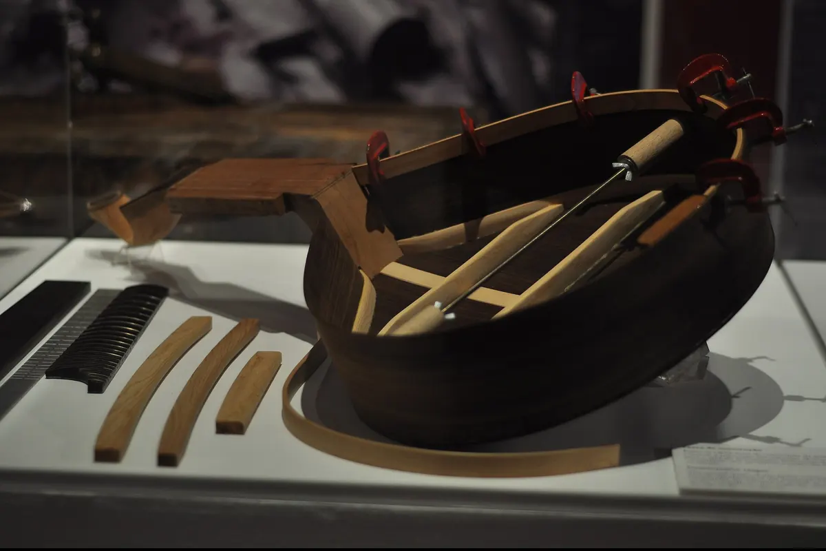 Il corpo incompiuto di una chitarra portoghese in una teca museale, con le doghe serrate e gli attrezzi del liutaio accanto.